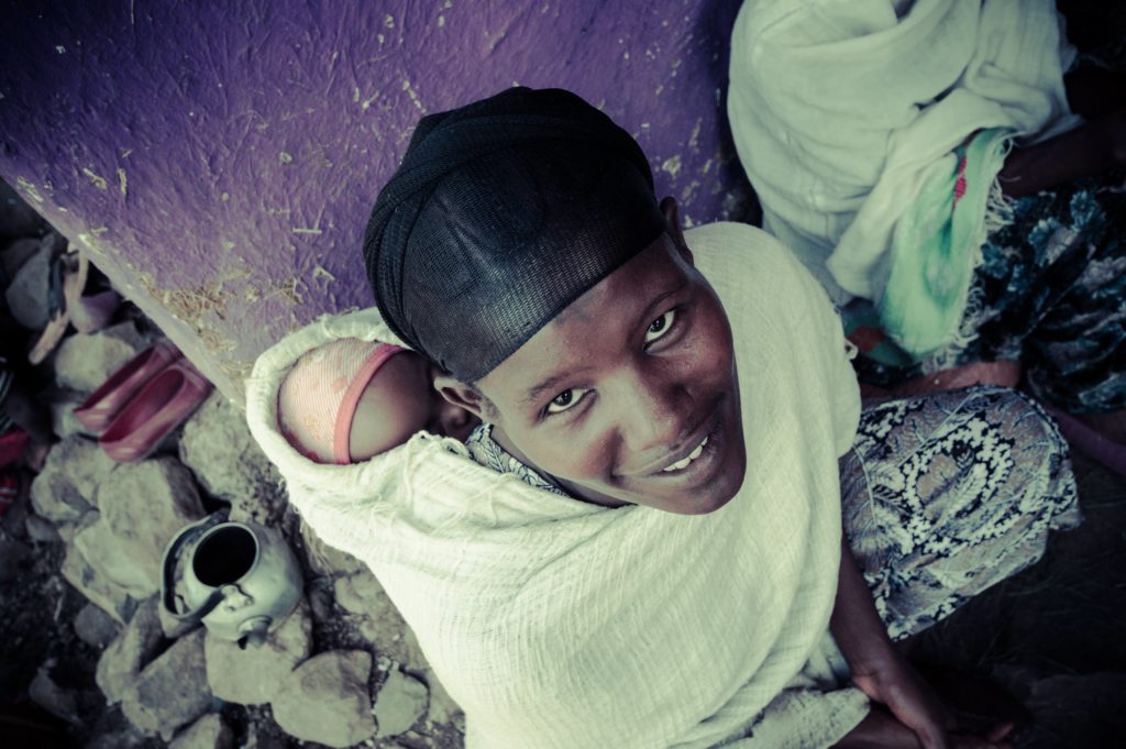 Mother with baby wrapped on back smiling. Photo by Warren Zelman.