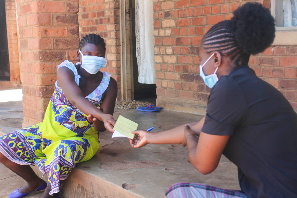 A community health volunteer speaks with a young woman sitting on the ground outside a brick building.