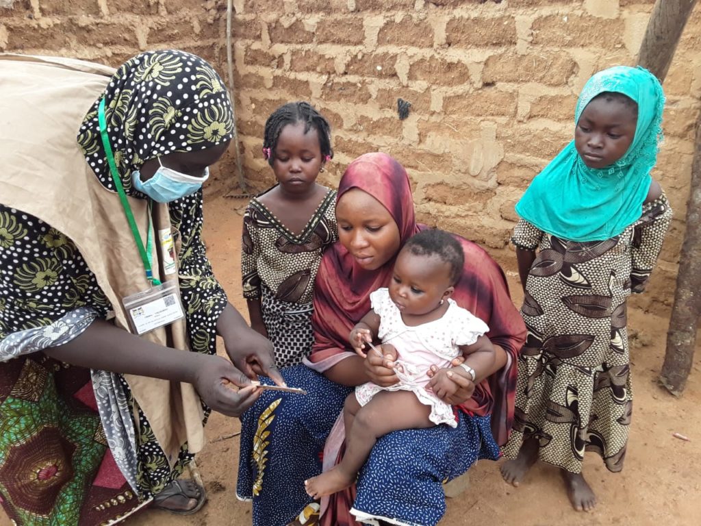 Una madre sentada con un niño pequeño en su regazo y sus otros hijos reunidos mientras un trabajador de la salud explica la importancia de la campaña SMC en Guene Alibori, Benin. Crédito de la foto Dra. Jocelyn Akakpo