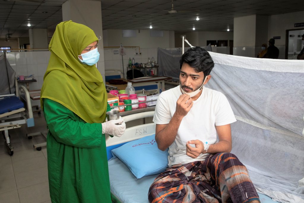 Um profissional de saúde vestido de verde-oliva com um lenço verde-oliva na cabeça administra medicamentos a um homem deitado em uma cama em uma unidade de saúde em Bangladesh
