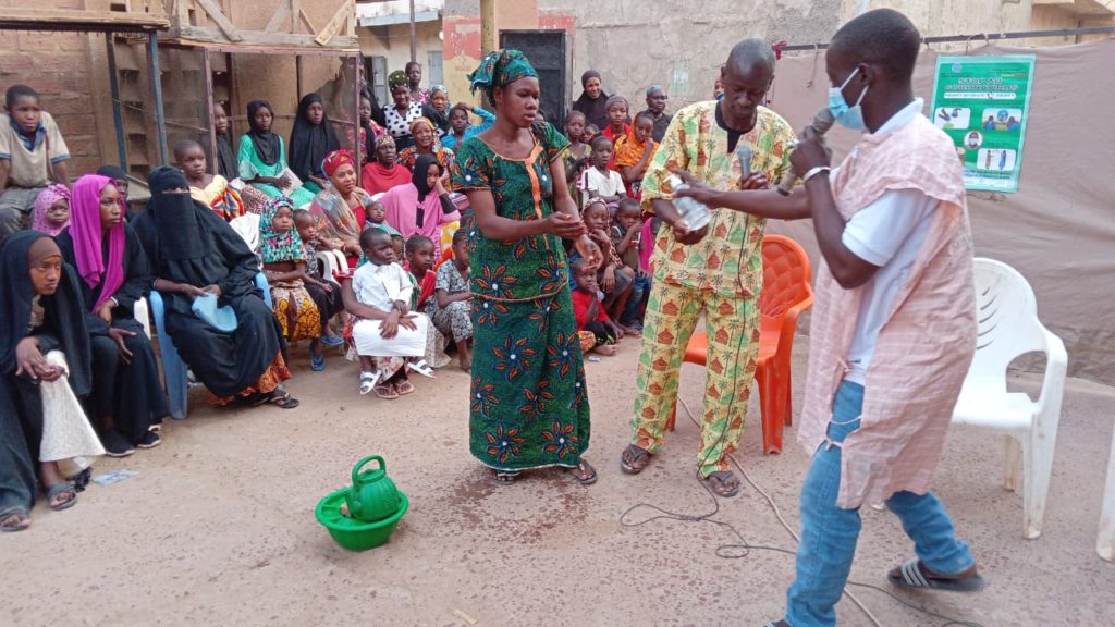In this image, people are gathered around to watch a theatrical presentation of two men and a woman acting out a scene as part of an educational training on sexual and reproductive health in Mali.