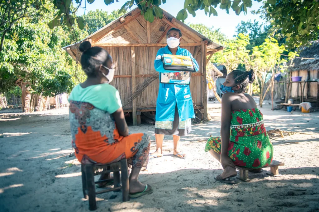 A community health worker in Madagascar stands in front of a women house in front of two seated women who have their backs to the camera, to conduct a health training. Photo credit: Samy Rakotoniaina