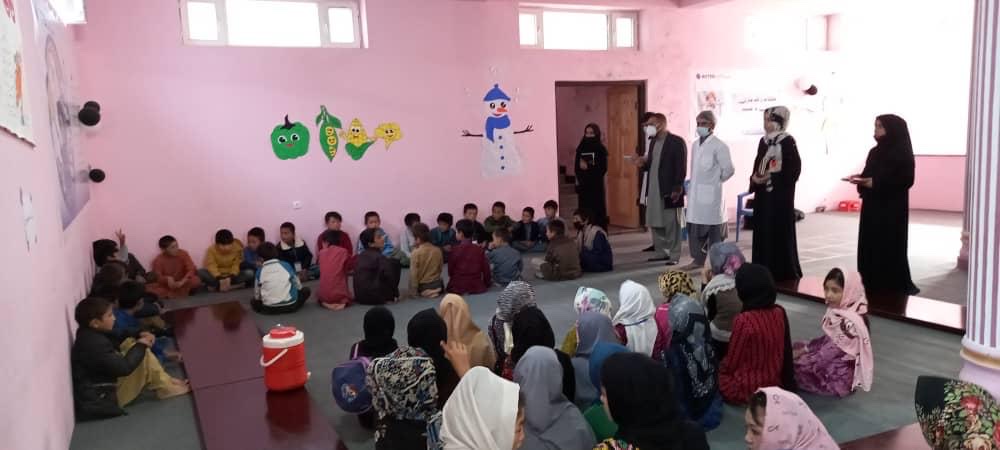 Foto de un salón de clases de estudiantes afganos de primaria, sentados en semicírculo en el piso de un salón de clases, frente a cuatro adultos parados al frente del salón.