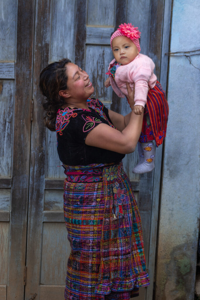 A Guatemalan woman dressed in traditional garb smiles as she holds her baby girl up in the air. The infant is dressed in pink with a pink cap adorned with a large pink flower.