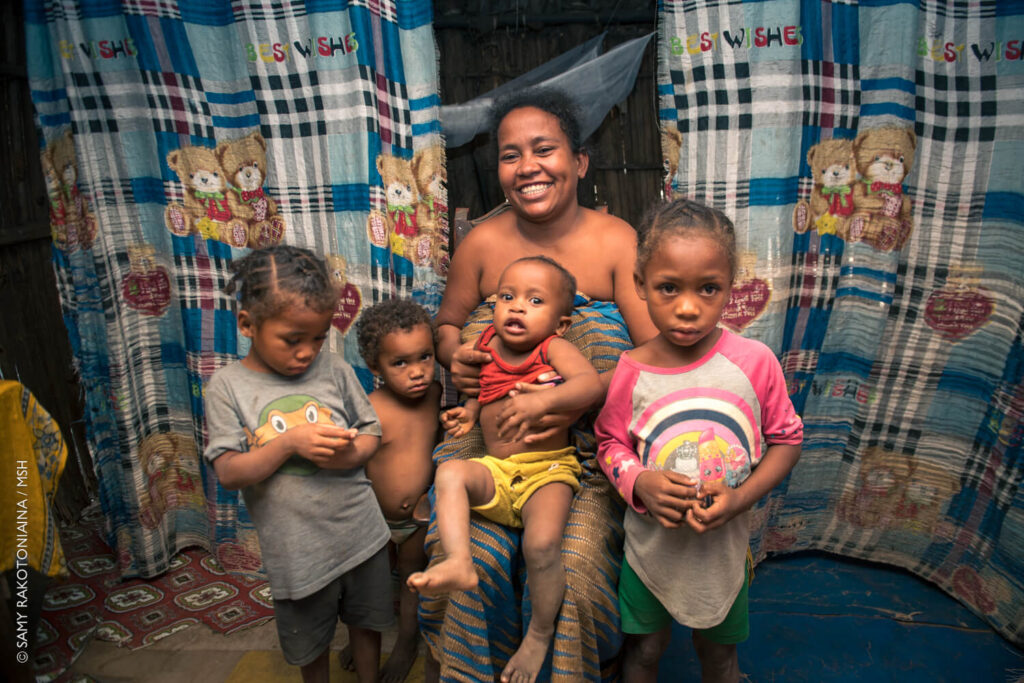 A mother in Madagascar poses with her for children while sitting on her bed, which has a mosquito net.