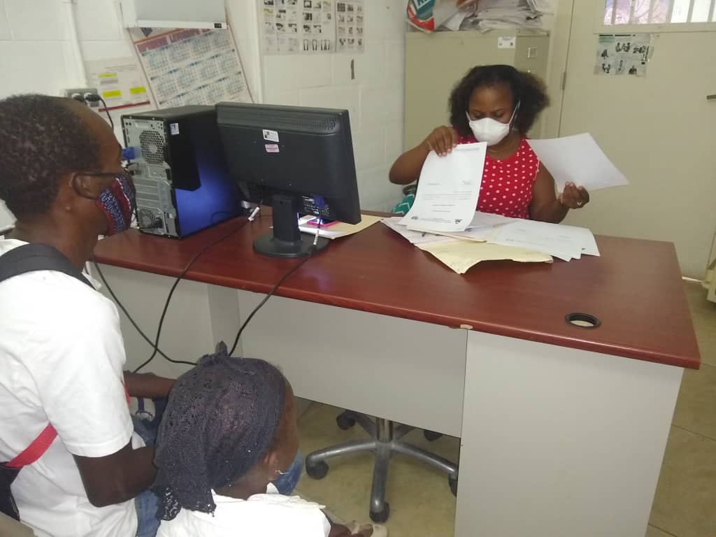 Uma enfermeira pediátrica clínica no Haiti usando uma máscara facial protetora senta-se em sua mesa examinando a papelada durante uma consulta de HIV. Dois clientes de costas para a câmera, sentados em cadeiras em frente à mesa.