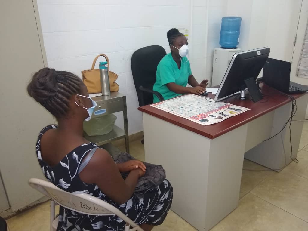 A nurse in Haiti wearing a protective face mask sits at her desk during an HIV case management consultation. A client with back her back to the camera, sit in a chair to the side. MOH staff for HLP/MSH