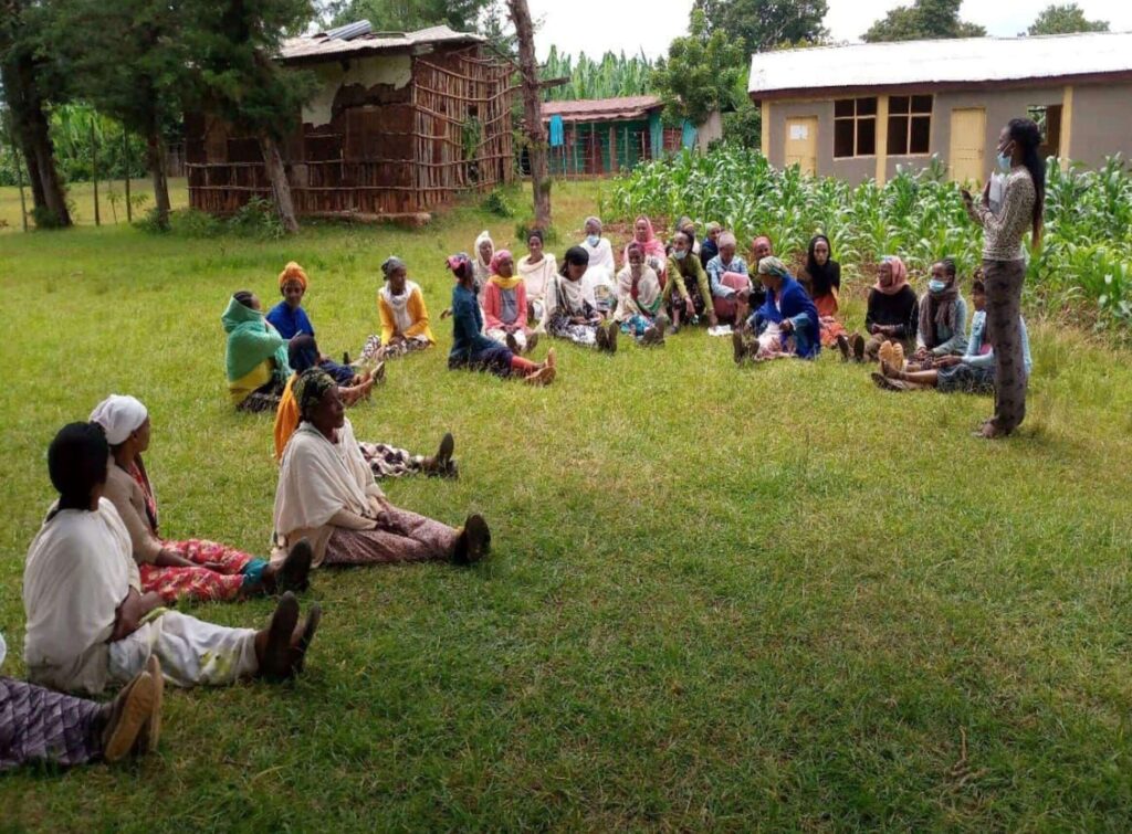 A group of people in Ethiopia are sitting in a semi circle on the grass in front of a brick building, listening to one person, who is standing, talk.