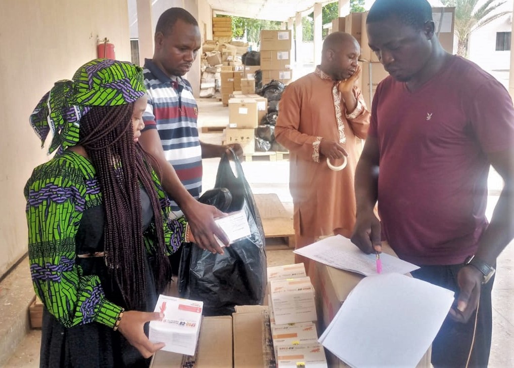 Four people in Nigeria - one woman and three men - are working outside on a porch under an awning packing malaria medicines into boxes and bags, and checking them off lists. Photo Credit: MSH