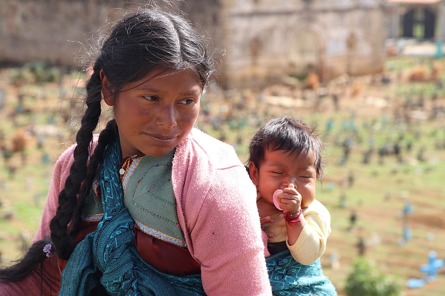 Il s'agit d'une image d'une jeune femme guatémaltèque, les cheveux tressés, regardant en arrière alors qu'elle marche avec un bébé attaché dans son dos à l'aide d'une écharpe traditionnelle.
