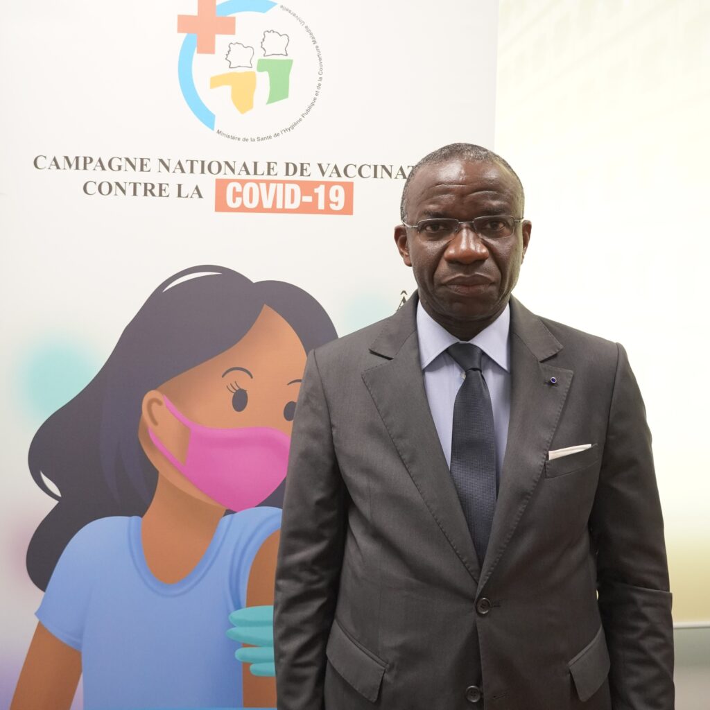 Doctor Raymond Brou, Technical Advisor to the Minister of Health, Côte d’Ivoire, standing in front of a advertisement for the country's COVID vaccination program.