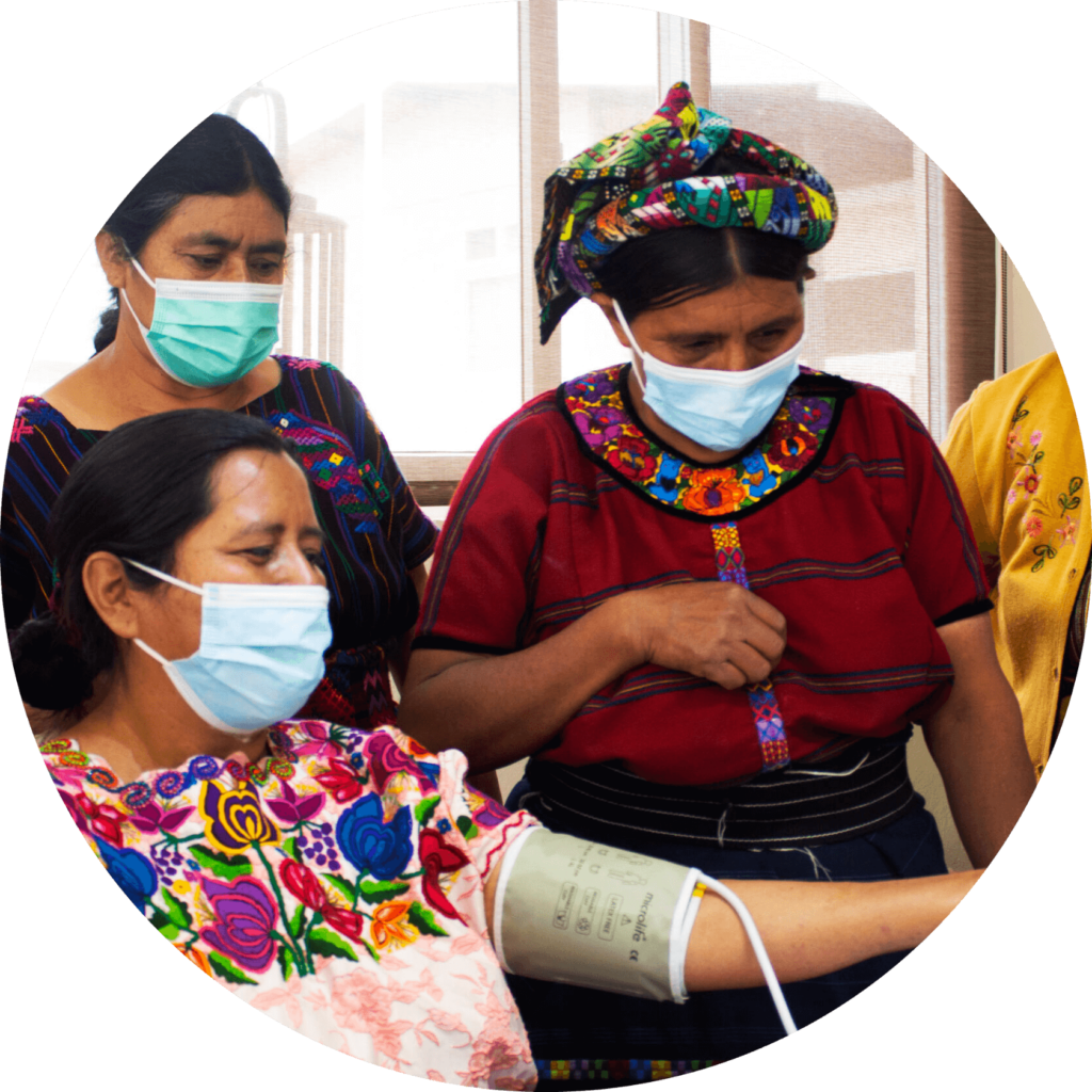 A health worker in Guatemala takes a woman's blood pressure, while two other women observe. All four women are in traditional dress.