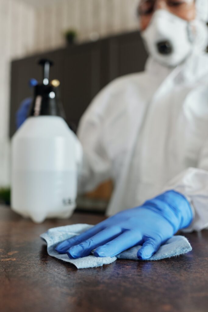 Bild eines Labortechnikers mit Maske und Schutzausrüstung, darunter blaue Gummihandschuhe, der eine Oberfläche abwischt.