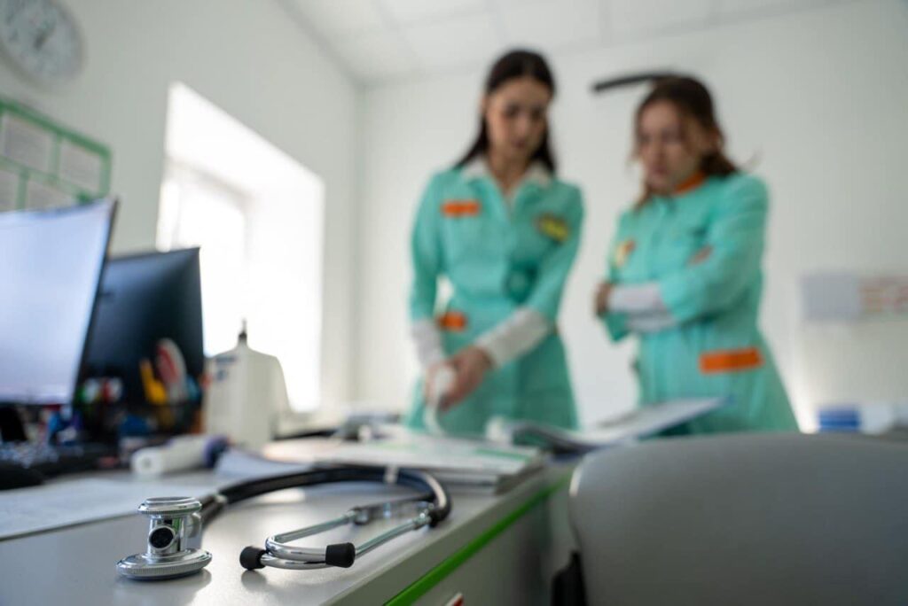 Two health workers in Ukraine in green uniforms look at a chart in the background, while the camera focuses on a stethoscope on a table top in the foreground.
