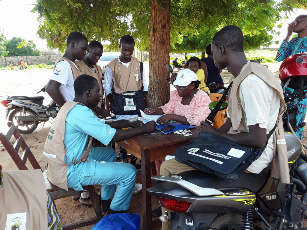 A team of field workers sit at a wooden desk in the shade of a large tree to go over paperwork