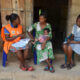 Nigerian community health worker Nneamaka sits in front of a cement brick home with a mother, also sitting, holding her infant, and another health worker, taking notes. Photo credit: U.S. President’s Malaria Initiative for States (PMI-S) activity
