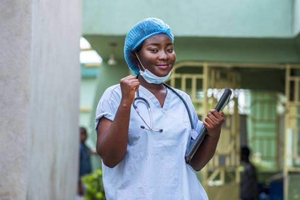 A healthcare worker wearing scrubs and a stethoscope stands outside a health facility.